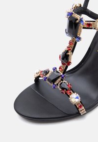 Μαύρο πέδιλο με ψηλό τακούνι και λουράκια διακοσμημένα με μεγάλα μαύρα, κόκκινα, μπλε και διάφανα πολύτιμα λίθους σε χρυσά πλαίσια.