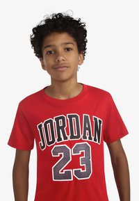 Maglietta rossa in cotone con la scritta "JORDAN" in lettere nere e il numero "23" in grigio, con scollo rotondo e maniche corte, texture liscia.