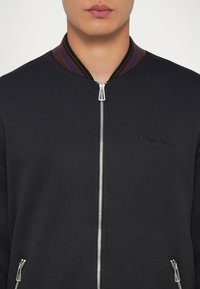 Schwarze Bomberjacke mit Reißverschluss vorne, strukturiertem Strickkragen mit lila und roten Akzenten sowie dezentem Logo-Stick auf der Brust.