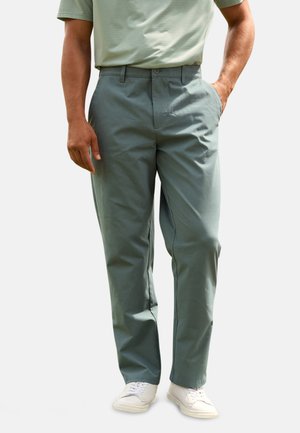 Hombre con pantalones casuales verde claro, zapatillas blancas y camisa de manga corta metida dentro, de pie con una mano en el bolsillo.