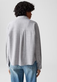 Persona con capelli ricci che indossa una maglietta a maniche lunghe grigia chiara e jeans blu sbiaditi, vista da dietro su uno sfondo semplice.