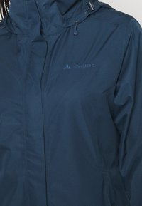 Veste imperméable bleu marine avec capuche et fermeture éclair, arborant le logo Vaude brodé sur la poitrine.