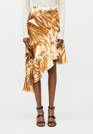 Nainen pukeutunut epäsymmetriseen beigeen ja ruskeaan lehtikuvioiseen hameeseen, jossa on röyhelöinen helma, yhdistettynä mustiin, remmikkäisiin korkokenkiin.