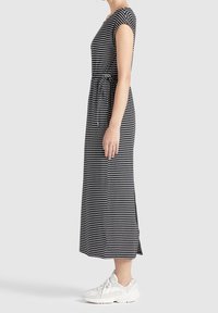 Kvinna som står sidledes i en svart och vit randig midiklänning med korta ärmar och vita sneakers.