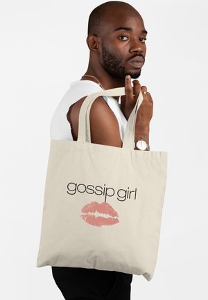 Osoba trzyma beżową torbę z napisem „gossip girl” i różowym odciskiem ust, nosi białą bluzkę bez rękawów i zegarek na lewym nadgarstku.