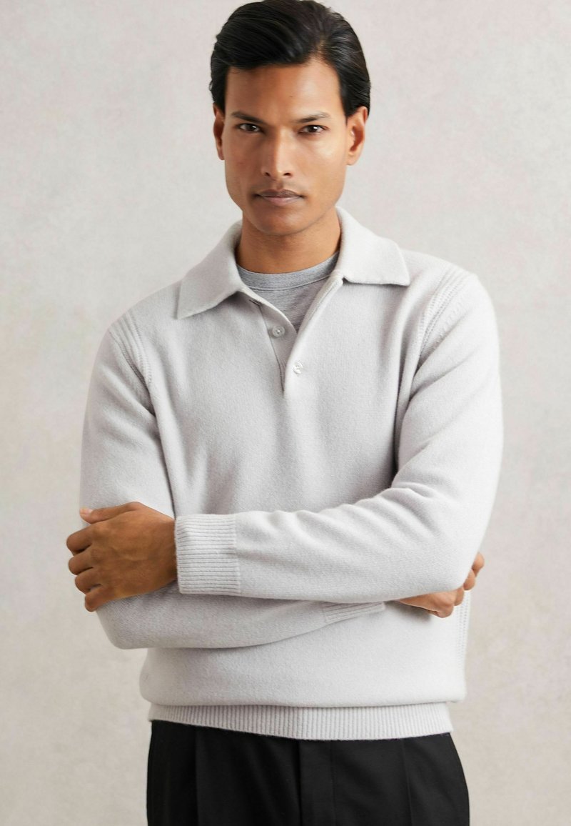 Pullover grigio chiaro con colletto, due bottoni e polsini a coste. Texture morbida con vestibilità rilassata. Abbinato a pantaloni neri.