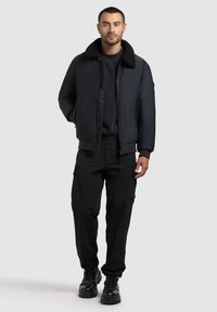 Musta pommijakku, jossa on pehmeä kaulus, päällä tumman sävyinen neule, yhdistettynä mustiin cargo-housuihin ja nauhallisiin saappaisiin. Yksinkertainen, käytännöllinen muotoilu.