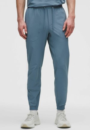 Mężczyzna w niebieskich joggerach z teksturowanej tkaniny, z elastycznymi mankietami i sznurkiem w talii, w białych trampkach i białych skarpetkach, stojący na białym tle.