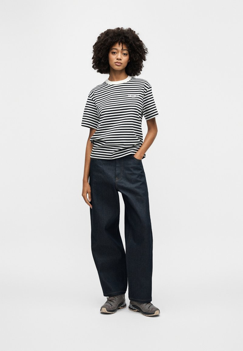 Kvinde med krøllet hår iført sort-hvid stribet t-shirt, vide mørke jeans og grå sneakers, står med den ene hånd i lommen.