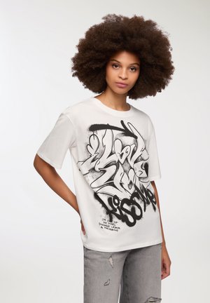 T-shirt bianco in cotone con grandi lettere e grafiche in stile graffiti nere. Maniche corte e scollatura rotonda. Jeans grigi usurati.