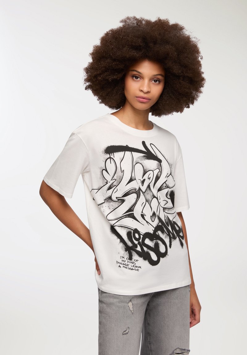 Biała bawełniana koszulka z dużymi, czarnymi napisami i grafikami w stylu graffiti. Krótkie rękawy i okrągły dekolt. Zniszczone szare dżinsy.