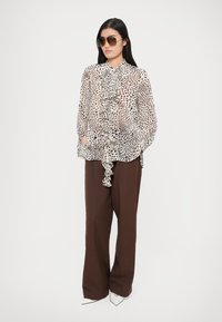 Bluză transparentă cu buline, cu volane în crem și negru, asortată cu pantaloni maro cu picioare largi și pantofi albi cu vârf ascuțit, accesorizată cu ochelari de soare.