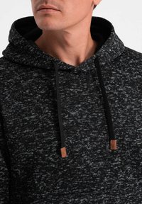 Muž nosící černou a šedou pletenou mikinu s kapucí a černými šňůrkami zakončenými hnědými koženými koncovkami.