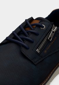 Chaussure en tissu bleu marine à surface texturée, accents en cuir marron contrastant, lacets bleu foncé et détail zippé sur le côté.