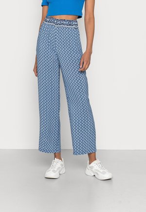 Mujer con pantalones anchos de tiro alto azules con patrón geométrico blanco, top corto azul y zapatillas blancas, de pie sobre suelo gris.