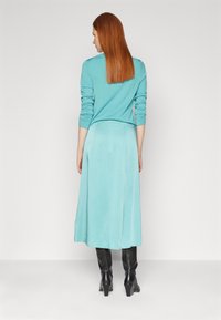 Un haut turquoise à manches longues associé à une jupe assortie, avec un design long et fluide. Des bottes noires à talons jusqu'aux genoux complètent la tenue.