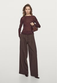 Langärmliges burgunderfarbenes Oberteil kombiniert mit weit geschnittenen dunkelbraunen Hosen. Das Model hält eine kleine braune Handtasche. Glattes Material mit figurbetonter Silhouette.