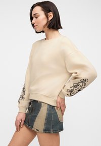 Γυναίκα με κοντά σκούρα μαλλιά φοράει μπεζ μακρυμάνικο φούτερ με κεντήματα λουλουδιών και μίνι φούστα από denim με μπλε και μπεζ συνδυασμό υφασμάτων.