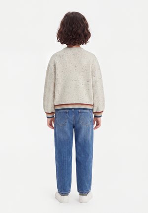 Enfant aux cheveux bruns bouclés, debout de dos, portant un pull beige tacheté avec des poignets rayés et un jean bleu avec des baskets blanches.