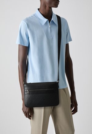 Black crossbody bag with two zippered compartments, leather material, geometric embossed pattern, worn over a light blue polo shirt.