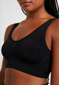 Svart bralette med sømløst design, brede stropper og et glatt, elastisk stoff med subtil innsamling i midten.