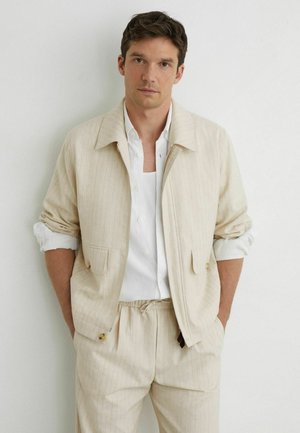 Mand iført beige jakke med nålestriber og matchende bukser med snøre, hvid skjorte under, hænderne i lommerne, stående op ad hvid væg.