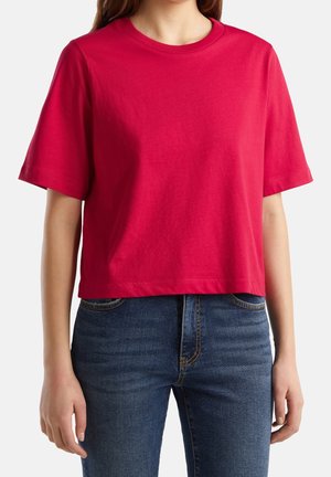 Kobieta w prostym czerwonym krótkim topie i niebieskich dżinsach, stojąca na białym tle.