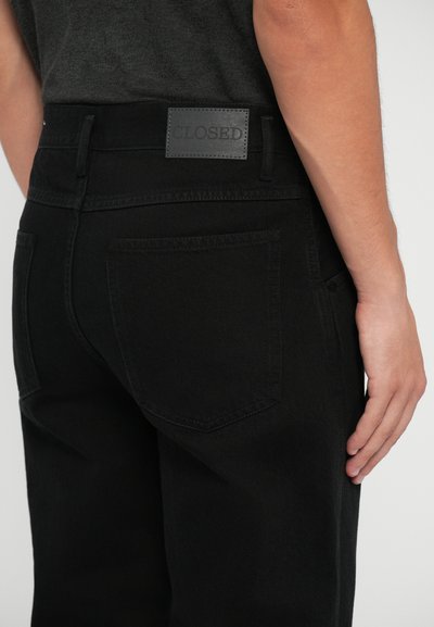 Pantaloni din denim negri cu o textură netedă, croi slim, buzunare la spate și o etichetă din piele rectangulară inscripționată "CLOSED" la talie.