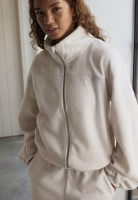 Femme portant une veste polaire crème avec une fermeture éclair à l'avant et un pantalon assorti, debout à l'intérieur près d'une fenêtre, les mains dans les poches.