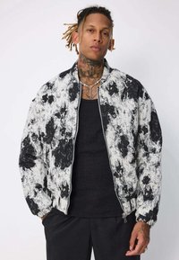 Veste bombardier noire et blanche avec un motif de projection abstrait, tissu texturé, poignets côtelés et fermeture éclair, associée à un débardeur noir.