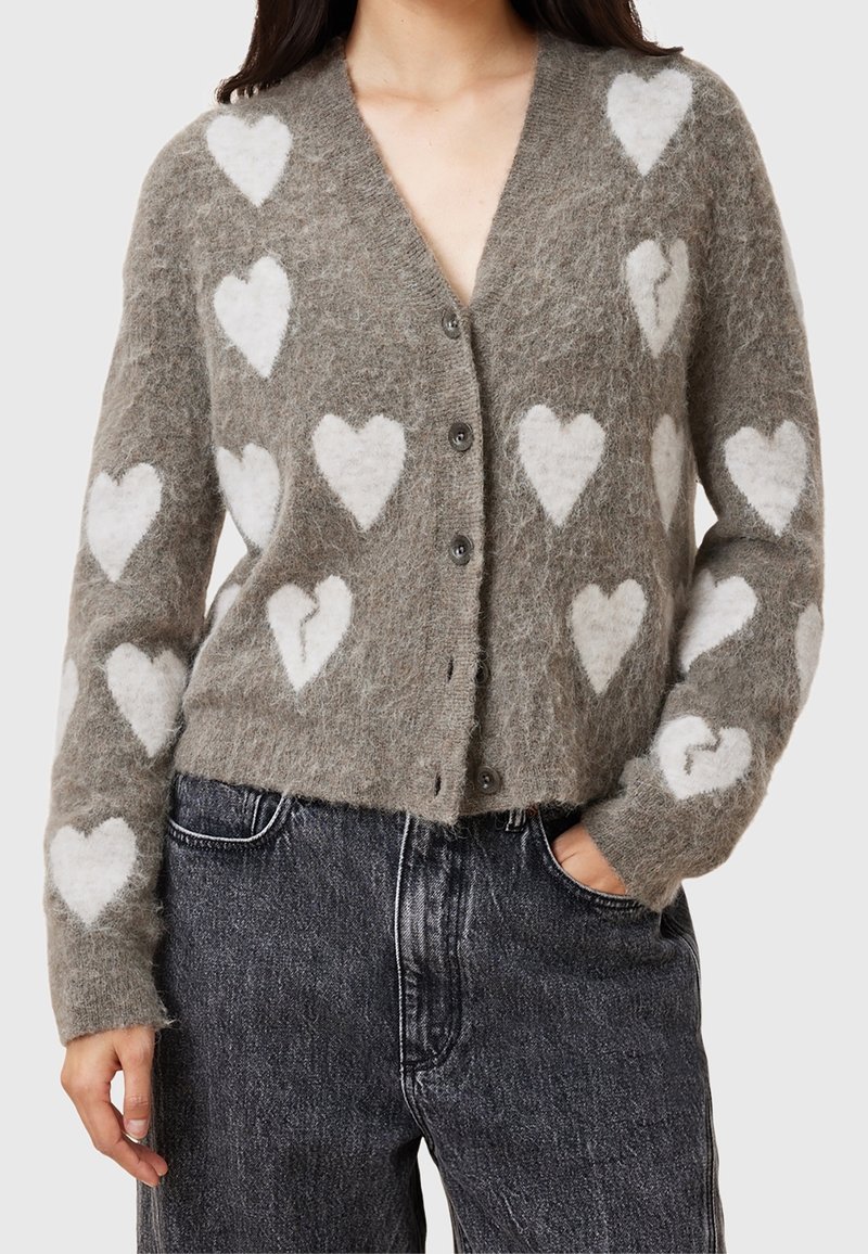 Femme portant un cardigan gris pelucheux avec des motifs de cœurs blancs, boutonné devant, associé à un jean noir taille haute, une main dans la poche.