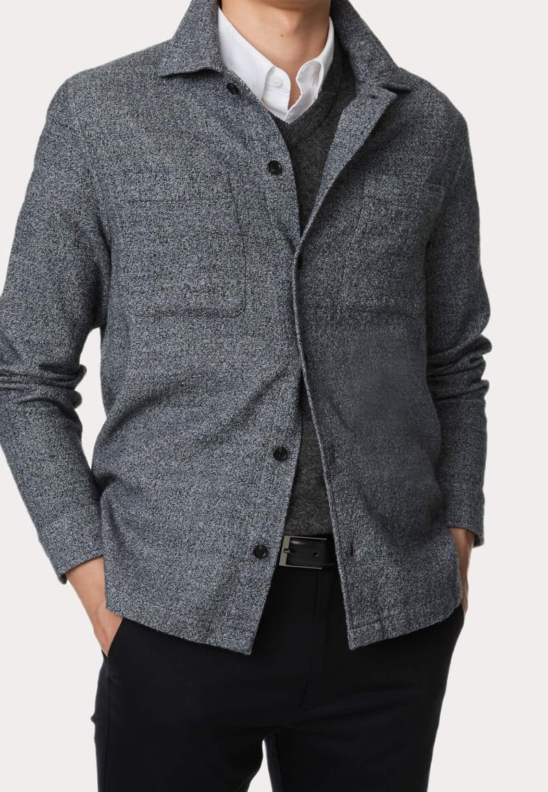 Veste-chemise grise texturée avec une fermeture à boutons, dotée de deux poches poitrine et d'un col, portée par-dessus un pull gris.