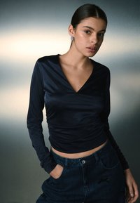 Marinblå långärmad topp med V-ringning, med en figurnära design och samlad textur. Matchas med högmidjade mörka denimjeans.