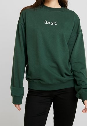 Femme portant un sweat-shirt vert foncé avec le mot BASIC brodé en blanc sur la poitrine et un pantalon noir sur un fond uni.