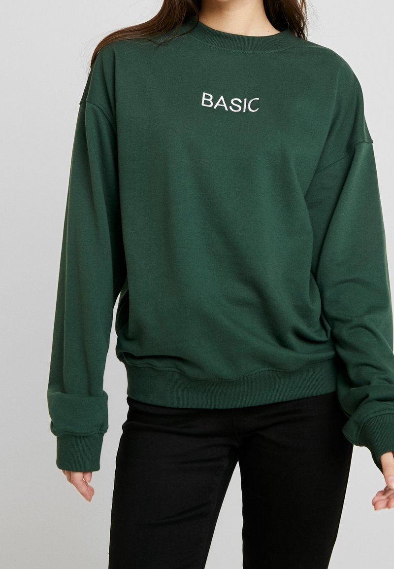 Femme portant un sweat-shirt vert foncé avec le mot BASIC brodé en blanc sur la poitrine et un pantalon noir sur un fond uni.