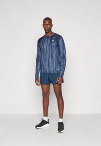 Camisola atlética de manga longa na cor azul-marinho com um padrão à risca, combinada com calções azul-marinho e ténis pretos. O tecido parece leve e respirável.