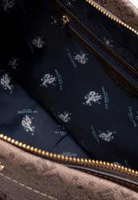 Brun mönstrad insida av en väska, som har en dragkedjeficka, textil med struktur och ett blått foder med tryckta logotyper och text. Guldfärgad dragkedjehardware synlig.