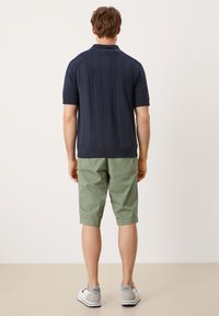 Marineblauw polo met korte mouwen en verticale ribbelstructuur, gecombineerd met lichtgroene kniebio shorts en grijze casual schoenen.