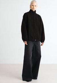Schwarze Fleece-Zipjacke mit hohem Kragen, lockerer Passform und elastischen Bündchen, kombiniert mit weit geschnittenen dunklen Denim-Jeans.