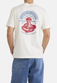 Camiseta de manga corta crema con un gráfico de serpiente roja y azul y texto en la parte posterior. Hecha de una tela suave, con un corte relajado.