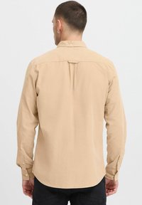 Beige hosszú ujjú ing, gallérral, gombos mandzsettákkal, textúrált anyagból, és egy hátsó redővel. Az alja ívelt.