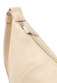 Krémová kožená taška s zakřiveným tvarem, obsahuje stříbrný zip a pravoúhlé upevnění popruhu. Hladký povrch, minimalistický design.