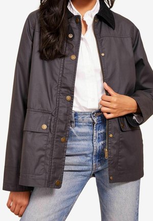 Kvinde iført mørkegrå jakke med knapper og lynlås over hvid skjorte og lyseblå jeans, med den ene hånd, der justerer lynlåsen på jakken.