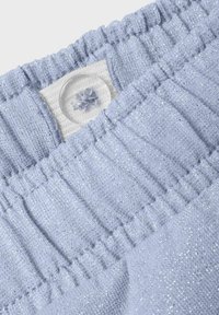 Tela texturada azul claro con un sutil brillo, que presenta una cintura elástica y un botón transparente que muestra un detalle de costura decorativa.