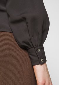 Jupe côtelée marron associée à un chemisier sombre en tissu lisse, doté de manches bouffantes et de poignets boutonnés.