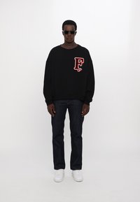 Zwarte oversized sweatshirt met een rood "F" logo op de borst, gecombineerd met donkerblauwe jeans en witte sneakers. Model draagt een zonnebril.