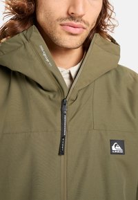 Olijfgroene hoodie met ritsluiting, voorzien van een logo patch en textiel dat extra warmte en duurzaamheid biedt.