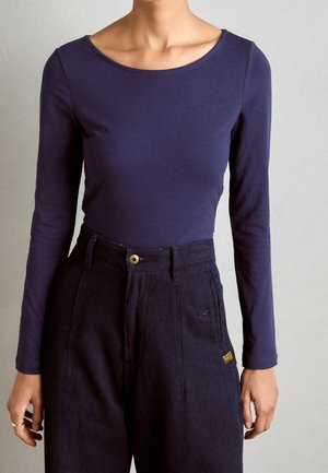 Personne portant un haut bleu marine ajusté à manches longues avec un col large et un pantalon bleu marine foncé taille haute avec poches et fermeture par bouton.