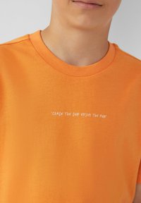 Oranje katoenen t-shirt met een ronde halslijn en korte mouwen. Het heeft een witte grafische tekst die zegt "LAAD DE ZON OP EN GENIET VAN DE PLEZIER."