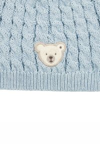 Cappello lavorato a maglia di colore azzurro chiaro, caratterizzato da un motivo a treccia testurizzata e una faccia di orso color crema ricamata sul davanti. Fondo a costine.